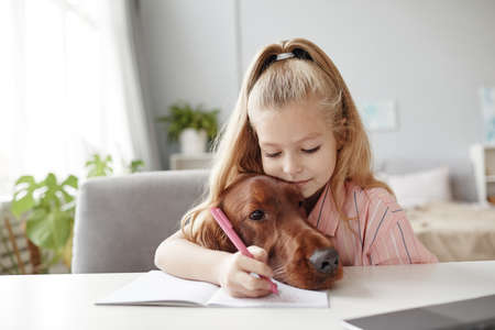 Portrait of cute blonde girl embracing dog while studying at home, copy spaceの写真素材