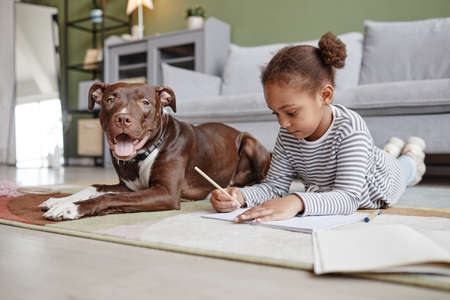 Full length portrait of cute African-American girl lying on floor with big dog, copy space