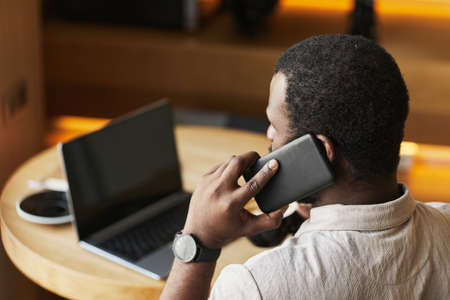 Back view portrait of adult black man speaking by smartphone while working at cafe table in wooden...