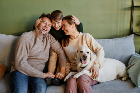 Portrait of happy family embracing son and dog while sitting on couch at home against green wall,...