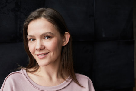 Close-up of young woman with long hair smiling at camera against the black wall