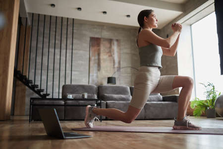 Young woman in sports clothing exercising on exercise mat in the room with online training on laptop