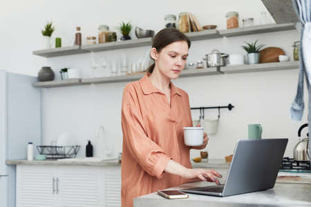 Young woman working online using her laptop while drinking coffee in the morning standing in the kitchenの写真素材