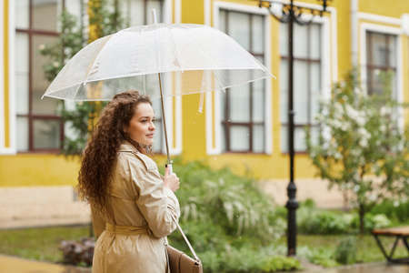 Horizontal shot of stylish young Caucasian woman with long curly hair standing outdoors under transparent umbrella on rainy day looking awayの写真素材