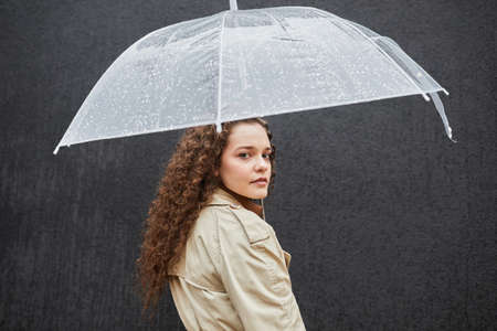 Horizontal medium shot of beautiful young woman with curly hair wearing trench coat standing under umbrella outdoors against dark gray wall looking at cameraの写真素材