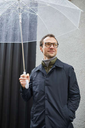 Vertical medium shot of stylish Caucasian man wearing eyeglasses standing outdoors under transparent...
