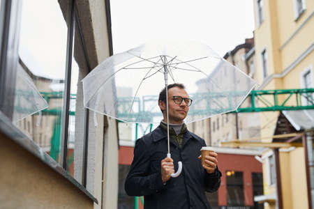 Horizontal medium shot of stylish Caucasian man wearing eyeglasses walking along street under...