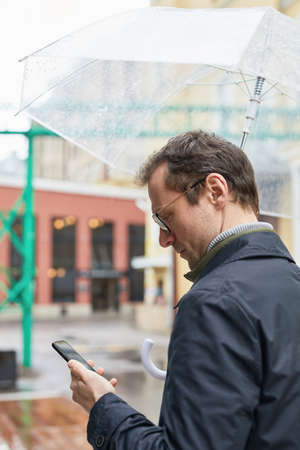 Vertical medium shot of modern Caucasian man wearing eyeglasses spending time outdoors standing under umbrella texting something on smartphoneの写真素材