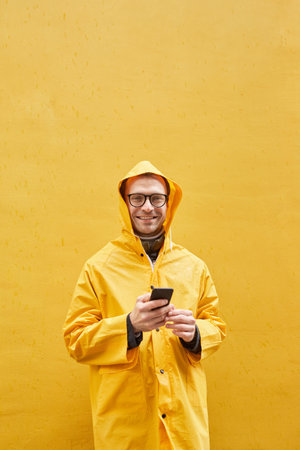Vertical portrait of modern Caucasian man wearing yellow raincoat standing against yellow wall holding smartphone looking at cameraの写真素材
