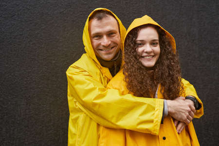 Horizontal portrait of Caucasian man and woman in love wearing yellow raincoats hugging and looking...