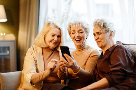 Group of three happy aged Caucasian women sitting together in living room against window and using smartphoneの写真素材