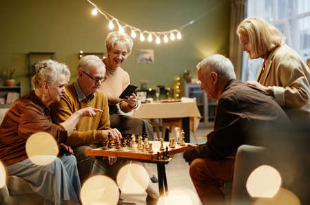 Three aged women watching two senior men playing chess during friends meeting in decorated living roomの写真素材