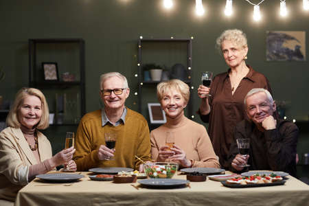 Group of five senior friends gathered at dining table, holding wine and champagne glasses while...
