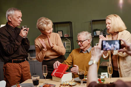 Joyful senior Caucasian man opening present box while sitting at table during birthday celebration...