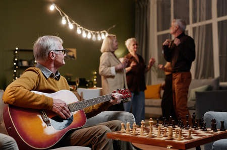 Old man sitting on armchair and playing acoustic guitar while his four friends dancing on background