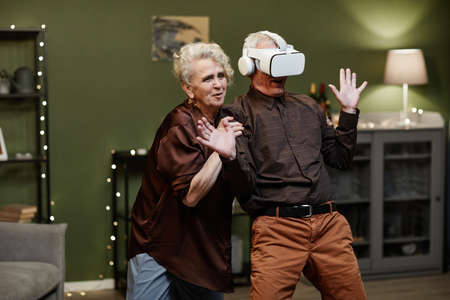Old Caucasian man standing in decorated with lights living room and enjoying modern VR glasses while...