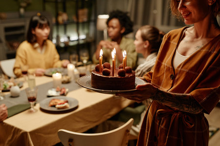 Young woman bringing birthday cake with candles for her friend during birthday party at homeの写真素材