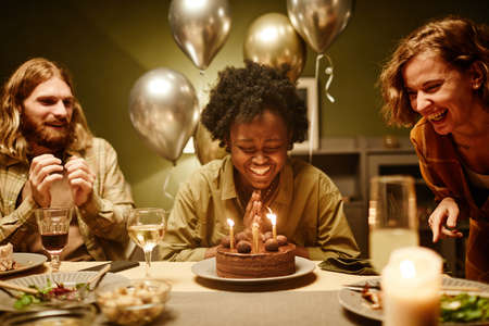 African happy woman sitting at the table with birthday cake with candles and laughing, she...