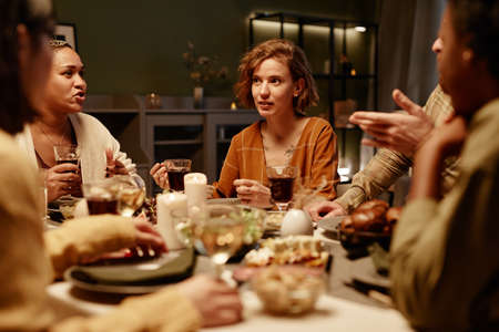 Group of friends sitting at dining table drinking wine and talking to each other at dinner party in...