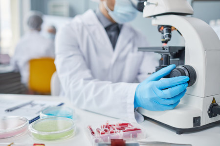 Close-up of scientist in lab coat and protective gloves examining samples with microscope while...