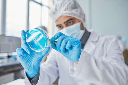 Young chemist in protective mask and gloves examining with blue liquid in tube in the laboratory
