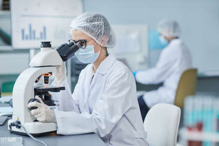 Young doctor in mask and lab coat looking through the microscope at the table, she examining medical...