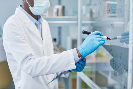 Close-up of chemist in lab coat and protective gloves writing formulas on glass wall while working...