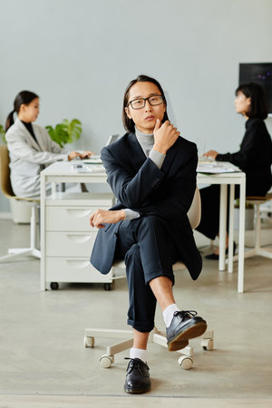 Minimal full length portrait of elegant Asian businessman looking at camera while posing sitting on chair in officeの写真素材
