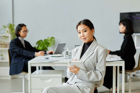 Minimal portrait of elegant Asian businesswoman looking at camera while taking notes in meetingの写真素材