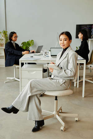 Minimal full length portrait of elegant Asian businesswoman looking at camera while sitting on chair in officeの写真素材