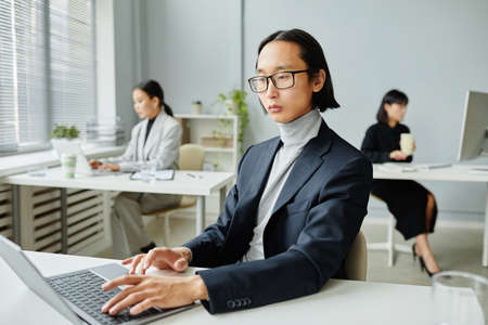 Minimal portrait of serious Asian man using laptop while working in office with female colleagues in backgroundの写真素材