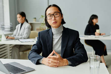 Front view portrait of Asian businessman looking at camera while sitting at desk in office with female colleagues in backgroundの写真素材