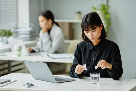 Portrait of young Asian woman dropping fizzing tablet in glass of water at workplace in office, headache and pain relief medicationの写真素材