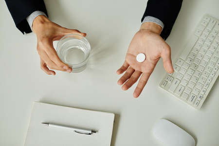 Minimal top view close up of unrecognizable businessman taking pill at white workplace in office,...