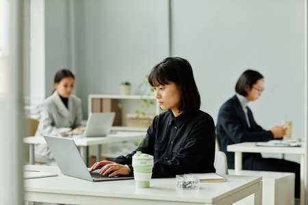 Side view portrait of young Asian woman looking at laptop screen while working in officeの写真素材