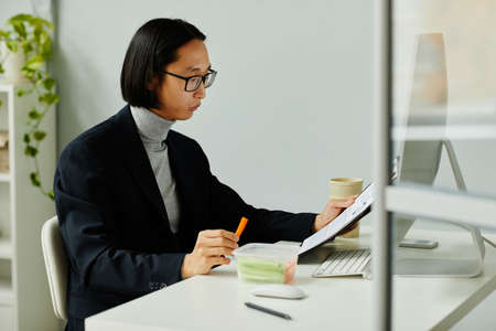 Side view portrait of Asian businessman eating healthy snacks while reading documents in officeの写真素材