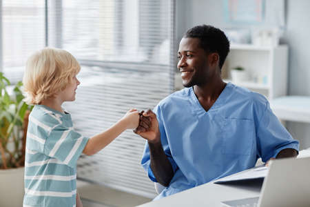 Side view portrait of smiling African-American doctor fist bumping kid during visit to pediatrician...
