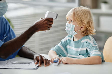Side view portrait of unrecognizable doctor measuring temperature of little boy during medical exam...