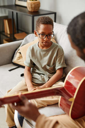 Vertical portrait of young African-American boy learning to play guitar at home with music tutorの写真素材