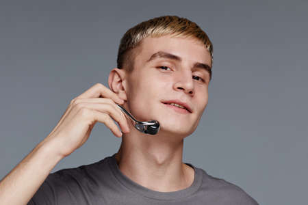Minimal portrait of blonde young man using face massager and smiling at camera against grey...