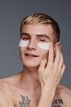 Vertical portrait of tattooed young man using moisturizing face patches and smiling at camera while enjoying male skincare routineの写真素材