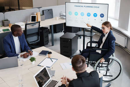 High angle portrait of young woman in wheelchair pointing at presentation screen while leading business meeting in officeの写真素材