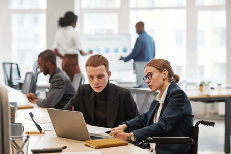 Side view portrait of young businesswoman using wheelchair while working with laptop in office, copy spaceの写真素材