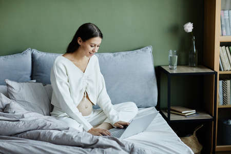Young pregnant woman typing on laptop and smiling, she communicating online sitting on her bed in...