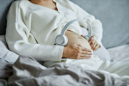 Close-up of pregnant woman putting on headphones on her belly and turning on the music while lying on the bedの写真素材