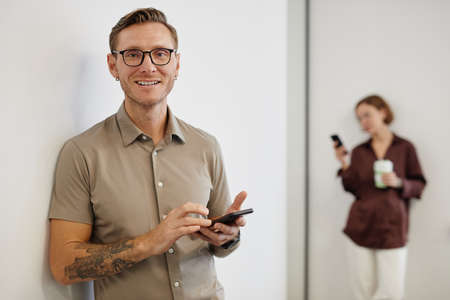 Minimal waist up portrait of smiling tattooed man holding smartphone and looking at camera in office, copy spaceの写真素材