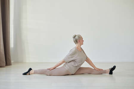 Side view of active elderly woman sitting on the floor in pose of hanumanasana while practicing yoga at leisureの写真素材
