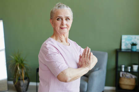Senior woman with grey hair keeping her hands put together against chest and looking at camera while...
