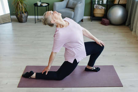 Elderly grey-haired woman looking upwards during yoga exercise while standing on mat and bending backwardsの写真素材
