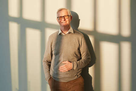 Smiling elderly man in brown pants and pullover and eyeglasses standing against sunlit wall with...
